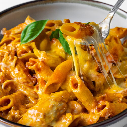A fork lifting a bite of cheesy 10-minute Instant Pot chicken chickpea pasta coated in tomato sauce from a shallow ceramic bowl, creating a long cheese pull with green basil leaves in the background.