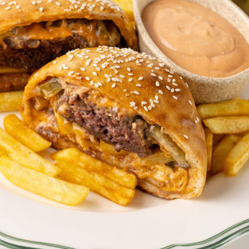 Two halves of a golden-brown baked stuffed burger bun filled with a juicy beef patty, cheese, and french fries, served on a white plate with french fries and a small bowl of creamy sauce.