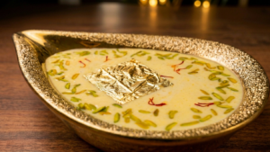Creamy Royal Almond Kheer garnished with sliced pistachios, saffron strands, and a square of edible gold leaf, served in an ornate gold teardrop bowl on a wooden table.