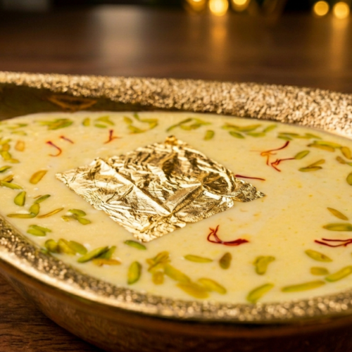 Creamy Royal Almond Kheer garnished with sliced pistachios, saffron strands, and a square of edible gold leaf, served in an ornate gold teardrop bowl on a wooden table.